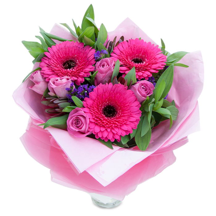 I picture this hand-tied arrangement as a snapshot of early spring in soft city light: three flamboyant fuchsia Gerbera daisies dominate the scene, their open faces and deep centres offering an immediate, joyful focal point. Around them, roses in a muted lavender-pink unfurl with layered, velvety petals that soften the overall energy and introduce a gentle, romantic scent that feels like a quiet promise. Sprigs of purple statice drift through the composition, their tiny clustered florets creating a delicate, airy texture and a complementary cool tone that plays beautifully against the warmer pinks. Verdant foliage-broad, glossy leaves akin to salal with hints of silvery eucalyptus-builds a generous, green foundation and adds a faint, leafy aroma. The bouquet is wrapped with intention: an inner cushion of pale pink matte tissue supports the stems while an outer veil of sheer, slightly crinkled fuchsia-pink cellophane gives a pretty shimmer and tidy finish; the top rim of a clear glass vase is visible at the base, suggesting the arrangement is already prepared for display. Emotionally, this design feels like an affectionate embrace-perfect for marking birthdays, Mother's Day, or a small anniversary-and suits the understated elegance of Belsize Village or a morning market stroll past the shops on Eton Avenue. The visual rhythm, scent, and textures reflect the care of a florist who chooses each stem to brighten someone's day.